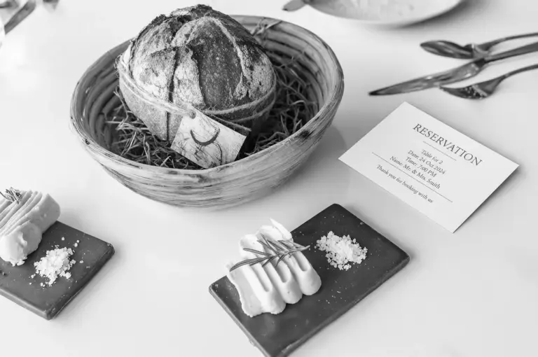 A rustic loaf of bread in a basket with hay, a reservation card nearby, and two rectangular plates with butter and salt on a white table setting.