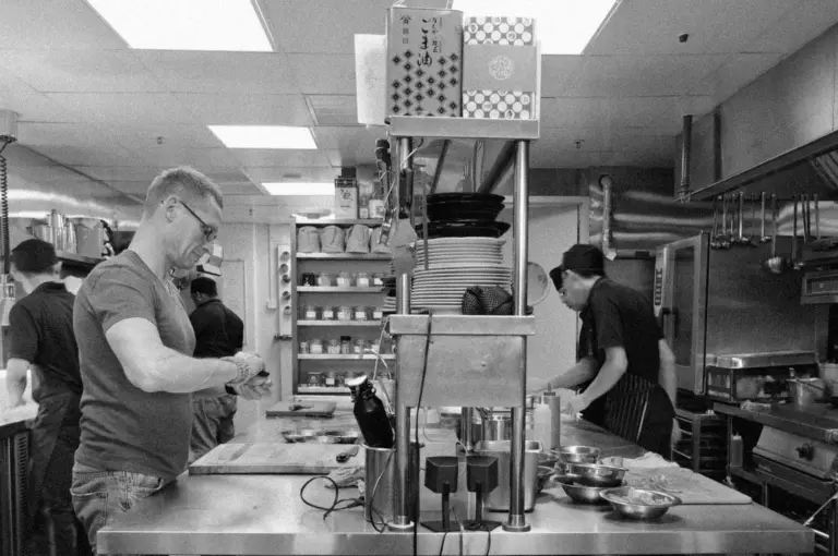 Black-and-white image of a busy restaurant kitchen with chefs working. One chef focuses on food prep, while others handle dishes. Plates are stacked on shelves.