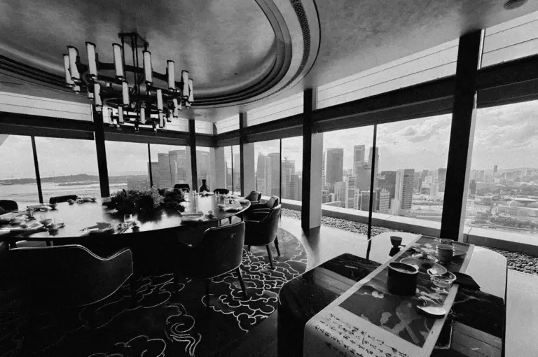 Luxurious dining room with a circular table, elegant chairs, and a modern chandelier. Floor-to-ceiling windows offer a stunning cityscape view. Black and white tones evoke sophistication.