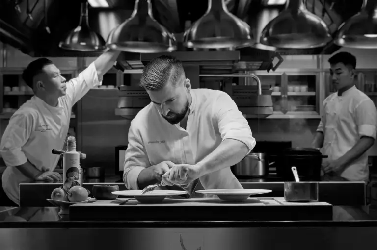 In a black-and-white kitchen, a chef attentively garnishes dishes, flanked by two assistants. The scene is framed by overhead lights, exuding focus and collaboration.