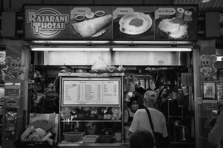 Black and white image of a street food stall named "Rajarani Thosai." Displays menu and food images of thosai, appom, puttu mayam. Customer stands in front