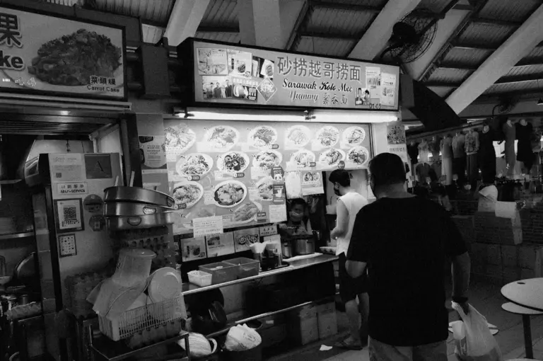Black and white image of a busy hawker stall with bright sign displaying menu photos. People are ordering, creating a lively, bustling atmosphere