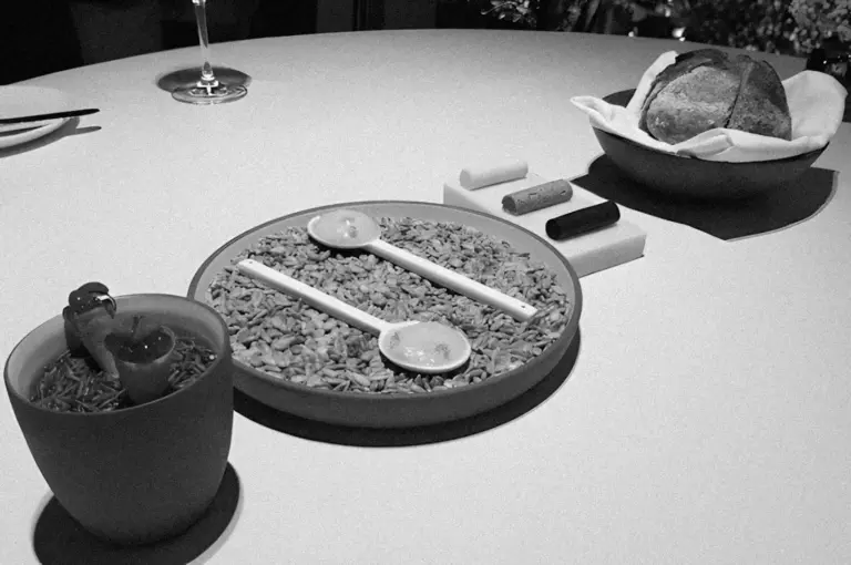 A monochrome image of a table setting with a bowl of risotto, two spoons, artistic breads, and a cocktail with a decorative figurine, creating an artistic and inviting dining atmosphere.