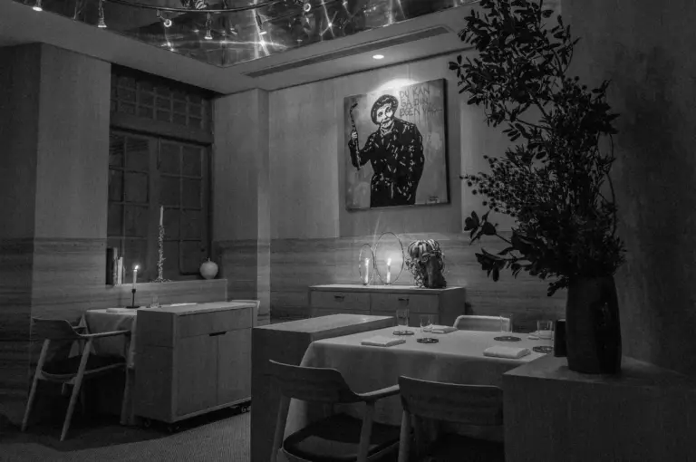 A black-and-white photo of an elegant dining room with modern decor. Tables are set with candles, a large painting hangs on the wall, and plants add warmth.