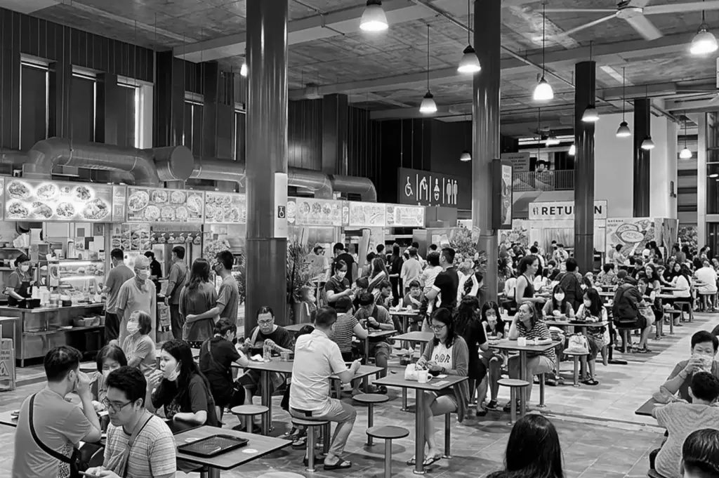 Black and white wide shot of a crowded indoor hawker centre with people dining at tables and food stalls in the background