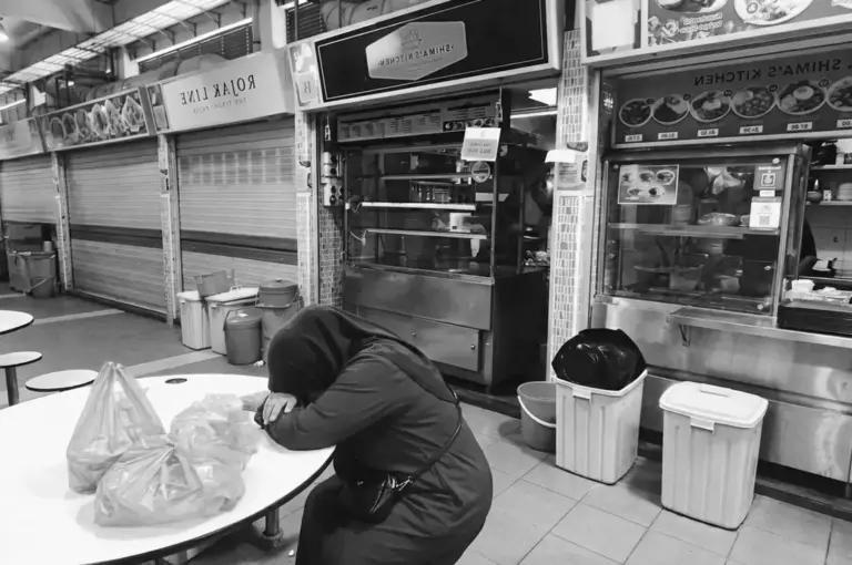 Exhausted hawker stall vendor resting head at table in food center with closed stalls behind