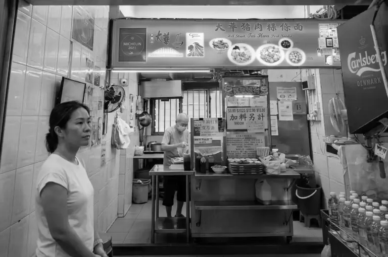 Hill Street Tai Hwa Pork Noodle stall interior showing Michelin 2021 recognition sign, menu pricing, and staff preparing food behind counter with customer waiting