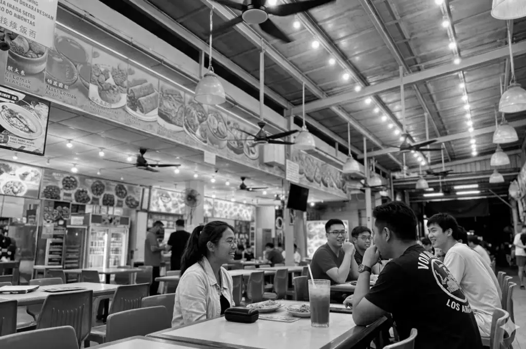 Singapore hawker center showing diners seated at tables, ceiling fans, pendant lights, and food stall menu boards displaying local dishes