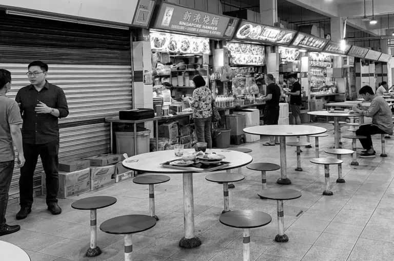 Black and white photo of a hawker center with round tables, patrons talking near open food stalls, and stacked boxes by a closed shutter
