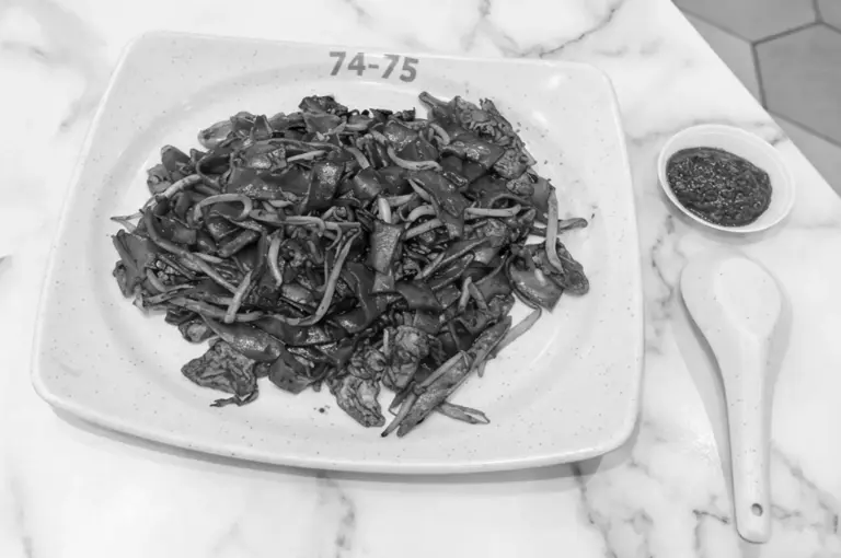 Black and white overhead shot of a plate of fried noodles (Char Kway Teow) with chili sauce on a marble table
