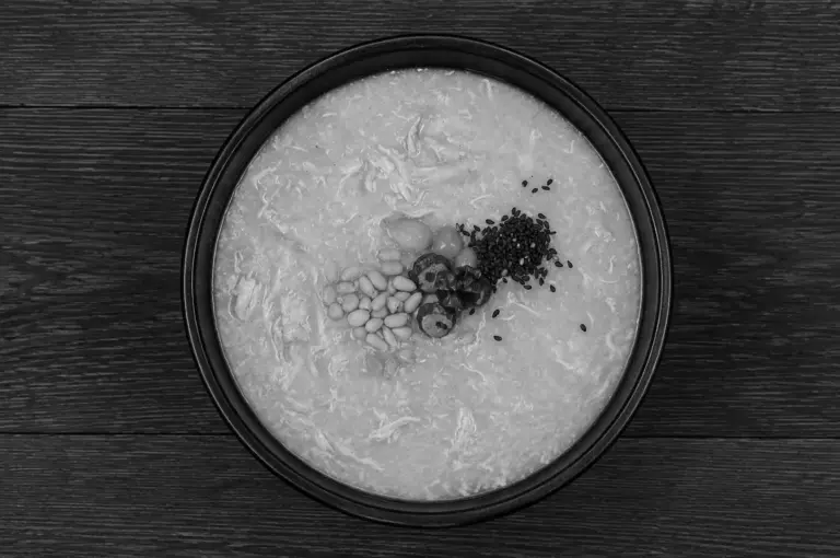 Black and white overhead view of samgyejuk (Korean ginseng chicken porridge) in a black bowl topped with pine nuts, black sesame seeds, and jujubes on wooden surface