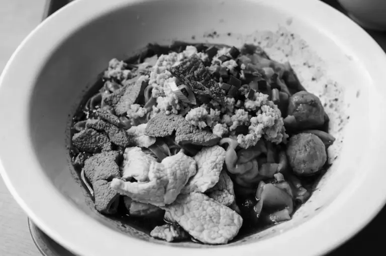 Close-up black and white image of a bowl of Tai Hwa pork noodle topped with minced pork, sliced pork, meatballs, and various toppings in dark sauce