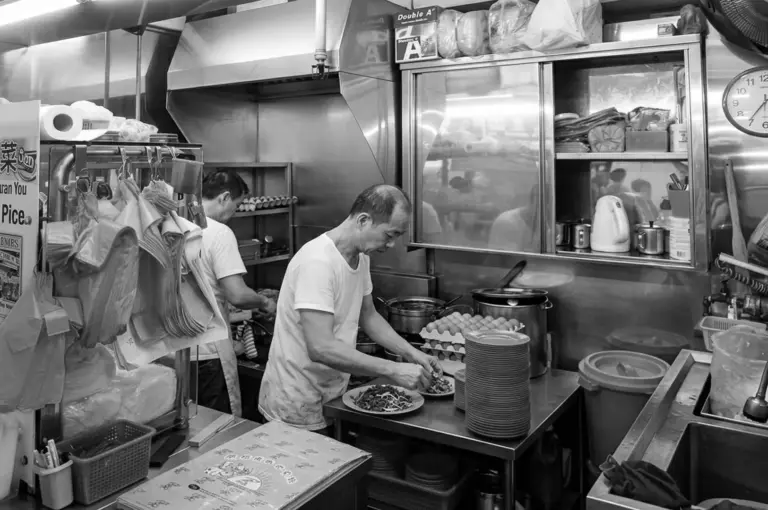 Black and white photo of hawker preparing plates of fried kway teow mee in the kitchen