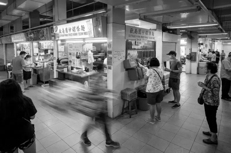Black and white wide shot of the busy Outram Park Fried Kway Teow Mee stall with customers waiting and walking by