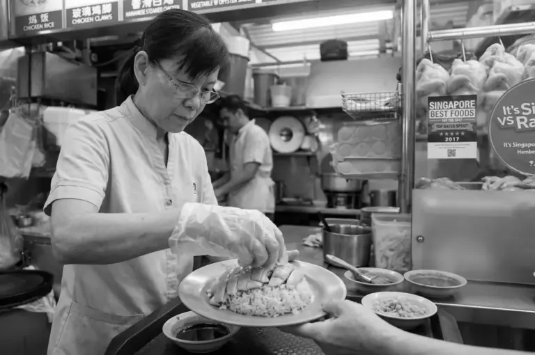 Tian Tian staff member in white uniform plating Hainanese chicken rice with sliced chicken over fragrant rice at the food stall