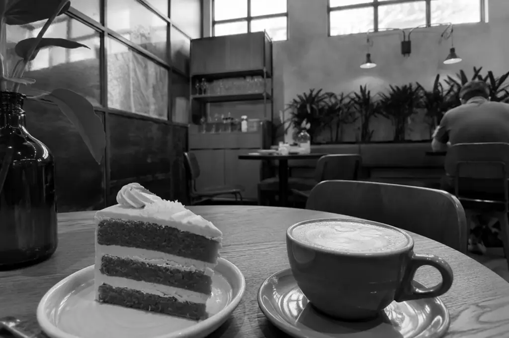 A slice of layered cake and a cup of coffee on a wooden table inside a cafe with large windows and plants