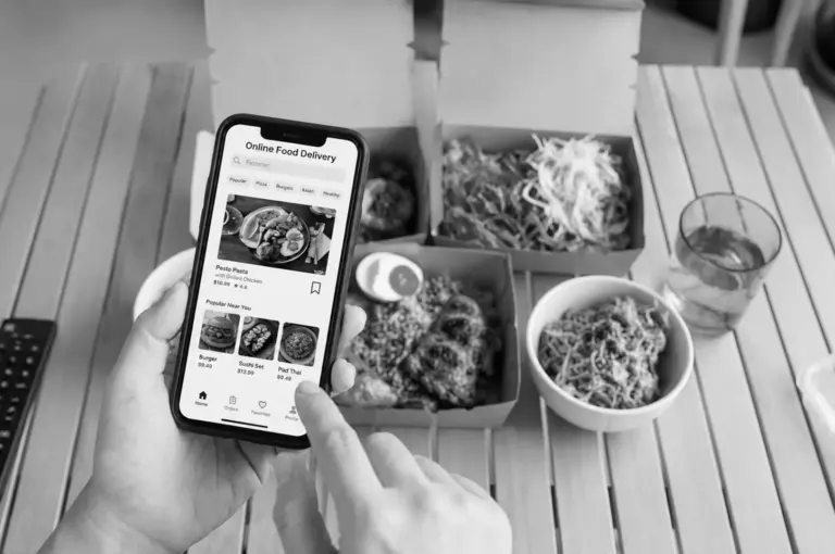 Person using food delivery app on smartphone with takeout boxes and noodles on a wooden table