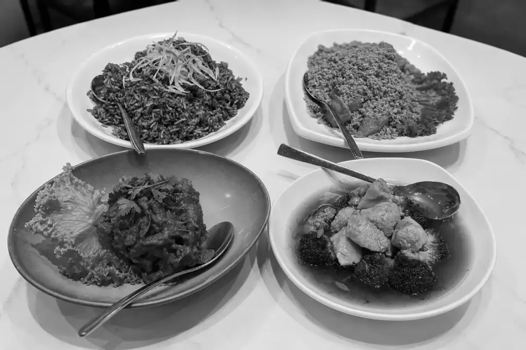 Four plates of vegetarian and vegan dishes including rice bowls, grain-based meals, and vegetable preparations with spoons on white marble table