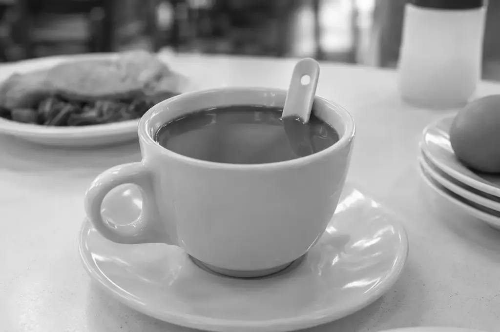 White ceramic cup of traditional Singaporean kopi with a ceramic spoon on a saucer, accompanied by soft-boiled eggs and a plate of food in the background