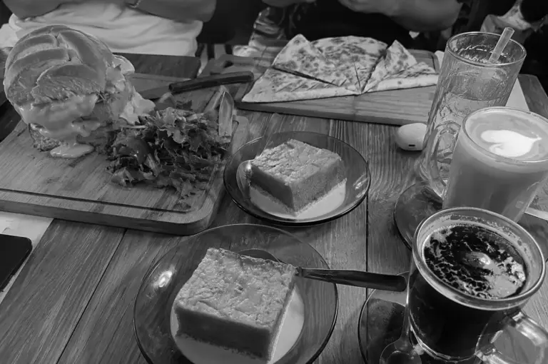Cafe table spread featuring a large burger, pizza slices, desserts, and coffee drinks on wooden tables