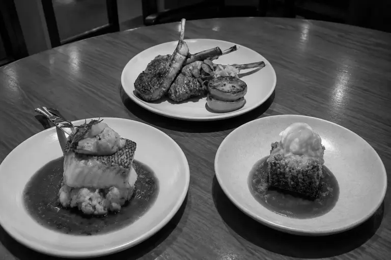 Three elegantly plated dishes including lamb chops, fish, and dessert are served on a wooden table at Bread Street Kitchen