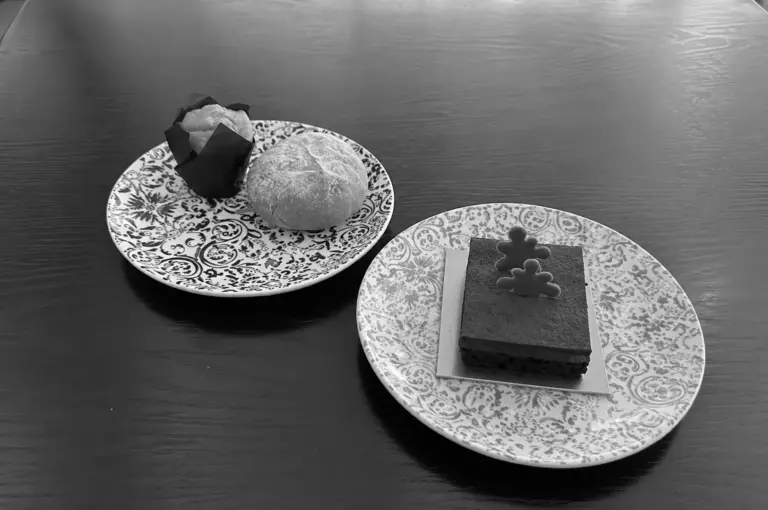 Black and white photo of high tea pastries on floral plates, including a bread roll and a chocolate cake slice with a puzzle piece decoration