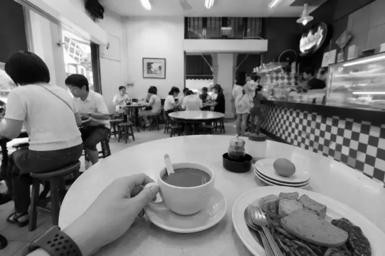 First-person view of a hand holding a kopi cup at a kopitiam table with soft-boiled eggs and a plate of noodles, with other diners and a checkered counter visible in the background