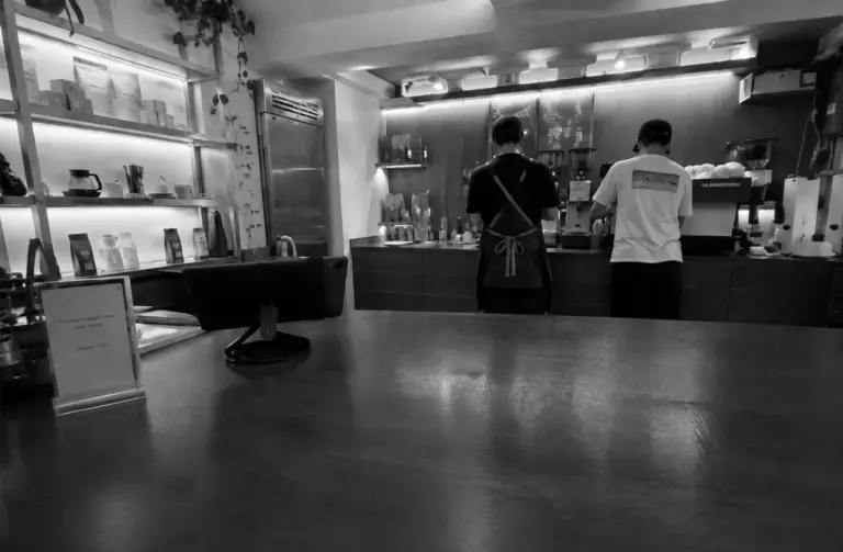 Two baristas prepare drinks behind the counter at Nylon Coffee Roasters, with coffee bags displayed on illuminated shelves