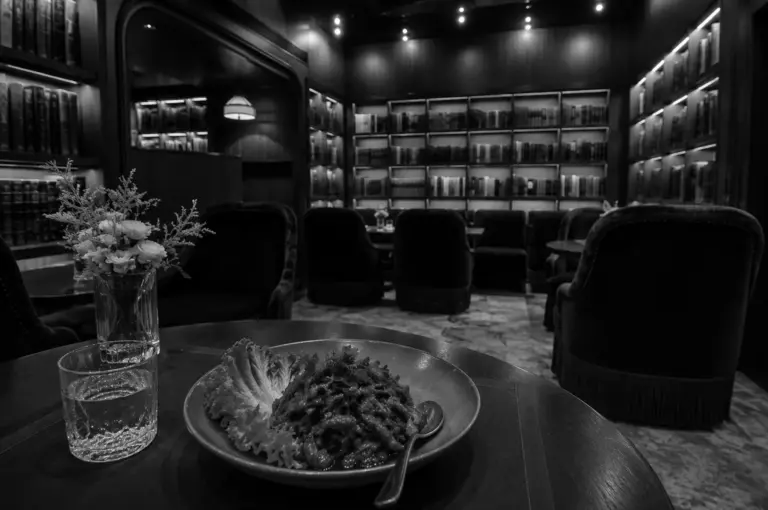 Elegant plant-based restaurant interior with bookshelf-lined walls, plush velvet chairs, and vegan dish plated on wooden table with water glass