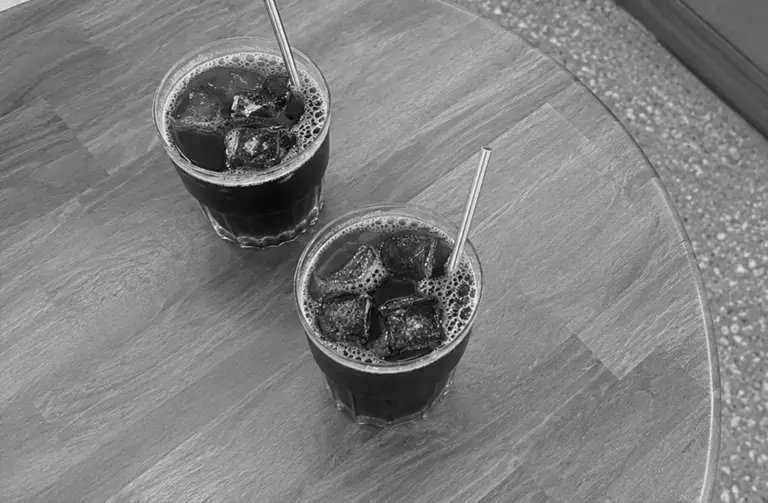 Two glasses of iced black coffee with metal straws sit on a round wooden table