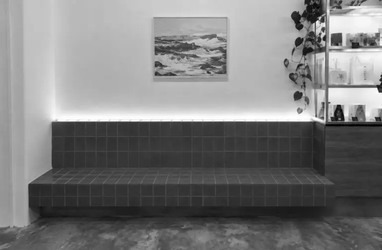 A tiled bench seating area sits beneath a framed ocean painting and trailing plants inside the minimalist cafe