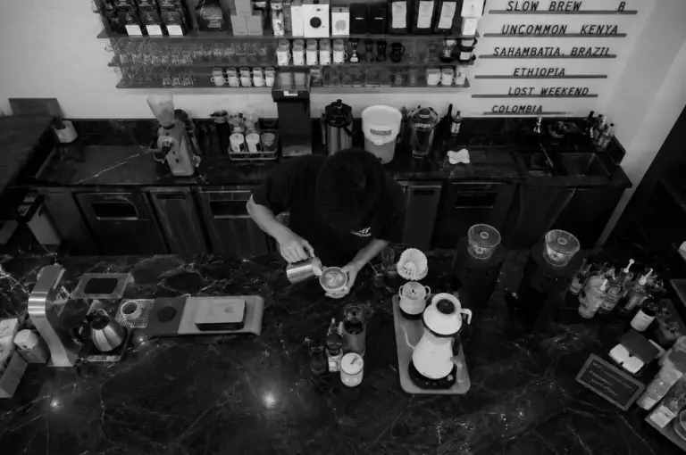 Top-down view of a barista pouring latte art at a marble counter with coffee bean shelves in the background