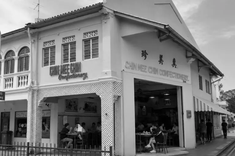 Street view of the Chin Mee Chin Confectionery corner building with vintage signage and patrons dining at tables along the covered walkway