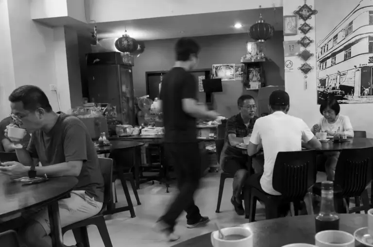 Patrons enjoying drinks at round tables in a traditional kopitiam with hanging lanterns and a staff member walking past