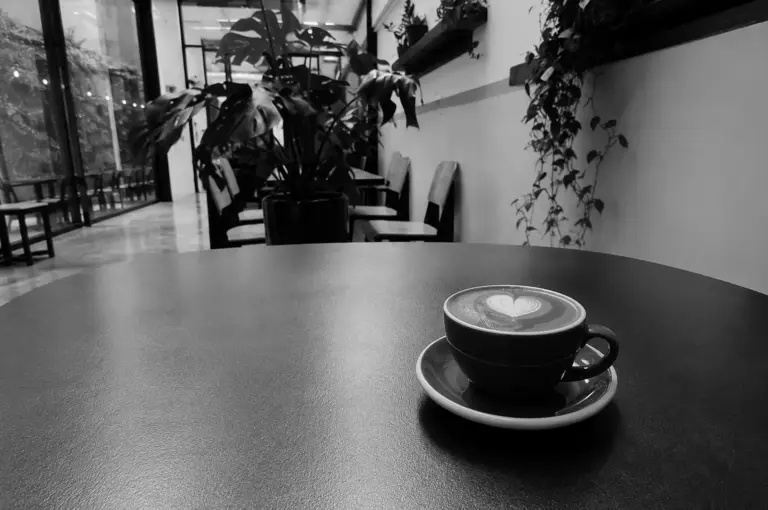 A cup of hot coffee featuring heart latte art on a dark round table, with a plant-filled cafe interior in the background