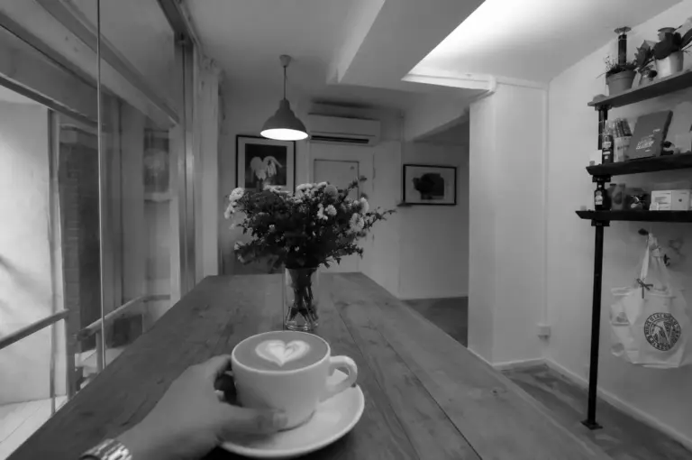 Hand holding a latte with heart art over a long wooden table with a flower vase in a cozy cafe interior
