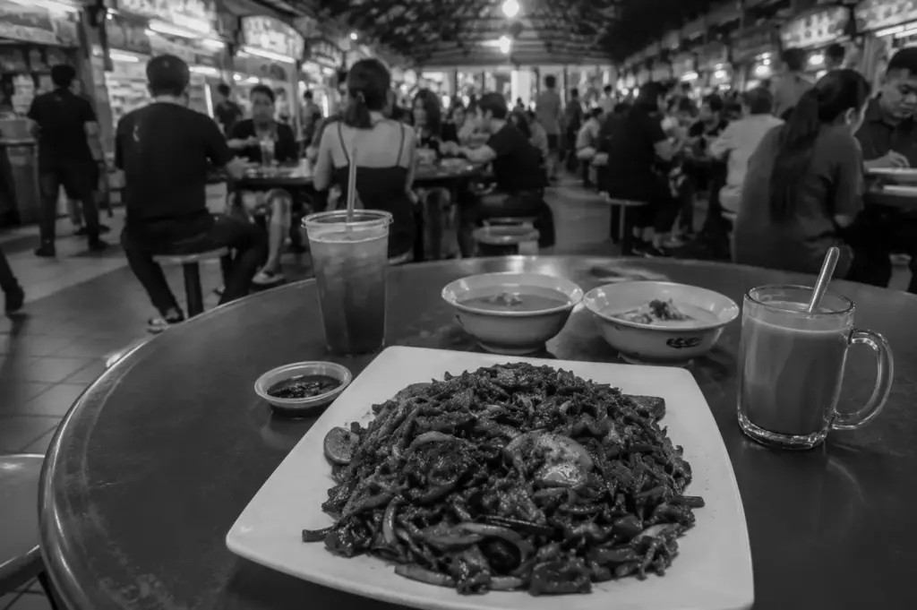 Plate of fried noodles with drinks on a table inside a busy hawker centre filled with diners