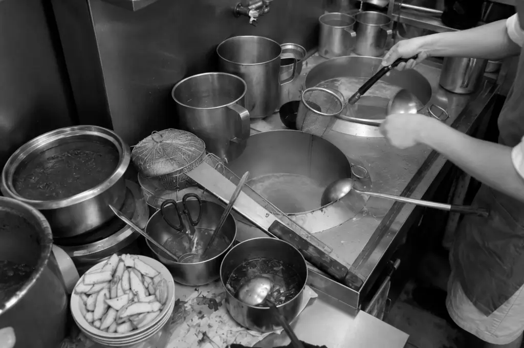 Young hawker preparing multiple pots of simmering broth in a busy kitchen, showcasing the hands-on work behind preserving Singapore's hawker food traditions.