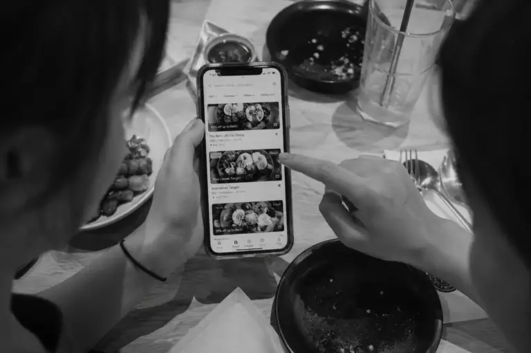 Black-and-white overhead view of two people browsing a food discovery app together on a smartphone at a dining table