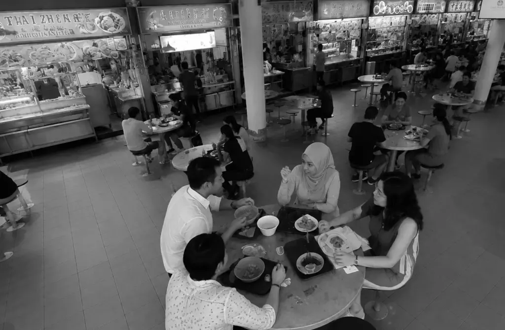 Hawker centre interior with diners seated among illuminated food stalls.