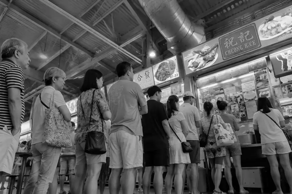 A line of customers waiting to order at a busy hawker centre roast meat stall beneath bright overhead signs.