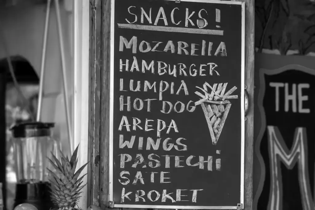 Chalkboard snack menu outside a restaurant listing items like mozzarella, hamburger, lumpia, hot dog, arepa, wings, and satay.