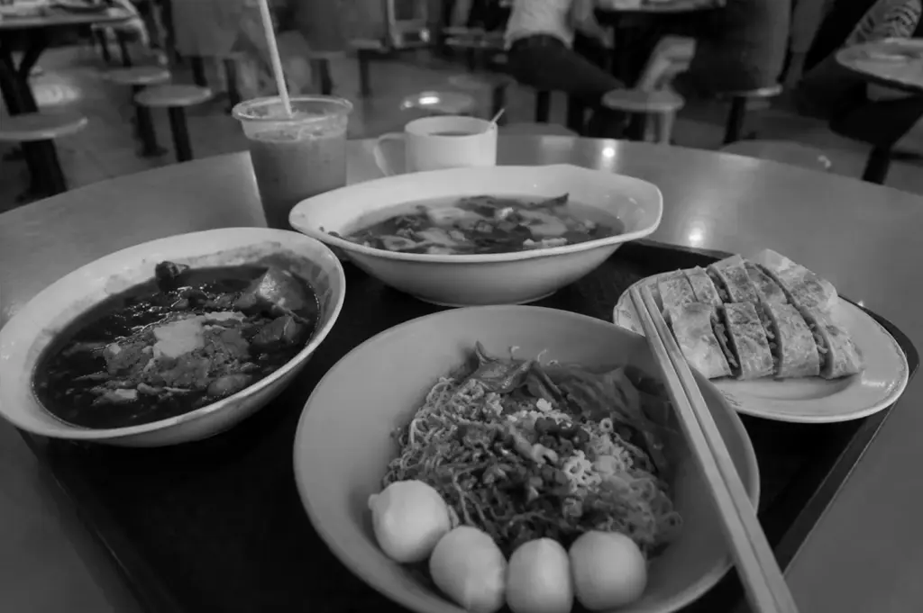 A nostalgic spread of hawker centre favourites, with noodles, soup, toast, coffee, and iced drink shared at a bustling communal table.