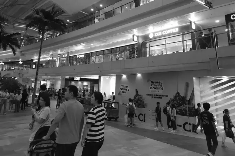 Busy multi-level shopping mall interior with shoppers walking past cafés and retail stores under bright lighting.