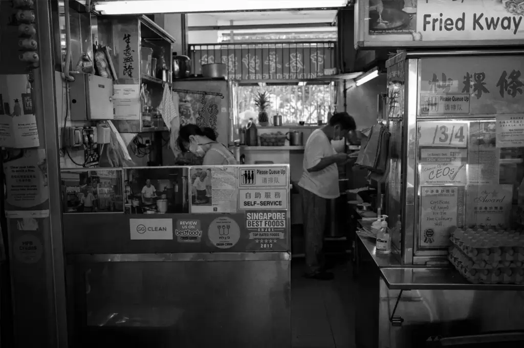 Inside a traditional hawker food stall, workers prepare meals behind a self-service counter decorated with awards and notices.