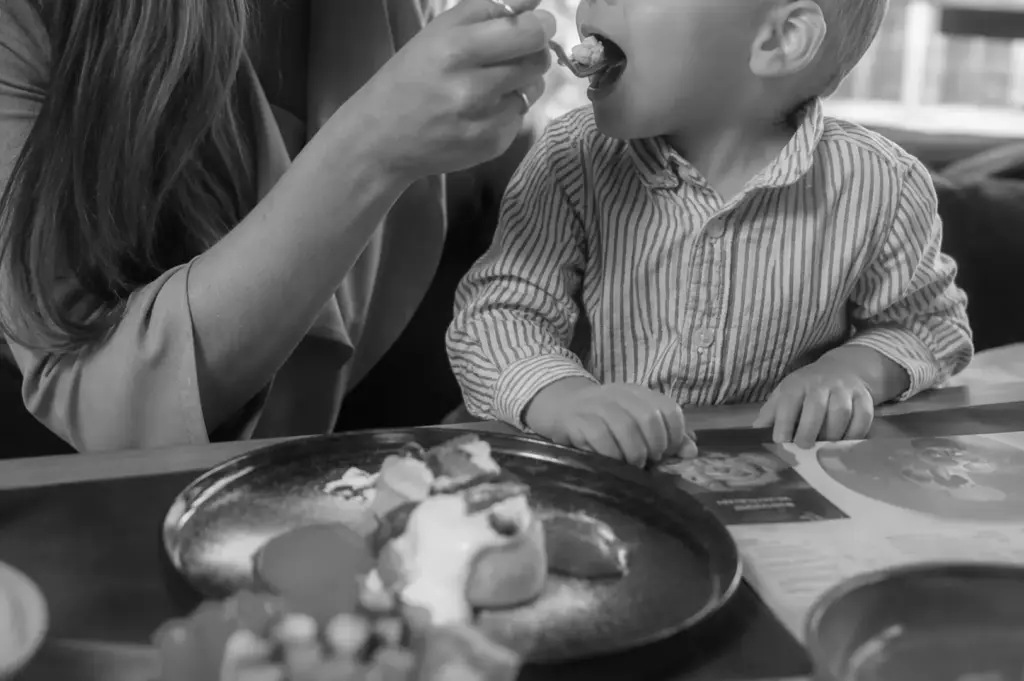 Adult feeding a young child with a spoon while sitting at a restaurant table with a plate of food.
