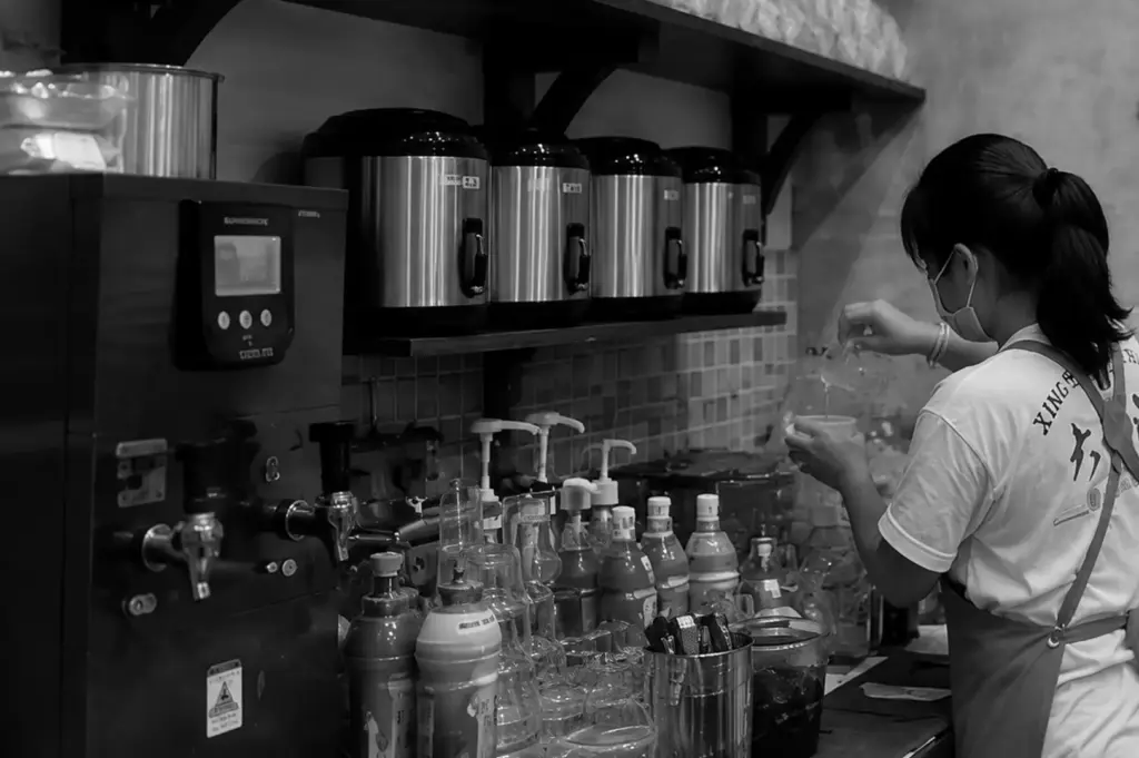 A hawker centre drink stall in action, where a vendor prepares beverages among rows of syrup bottles and drink dispensers.