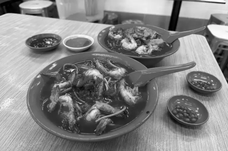 Two bowls of rich prawn noodle soup with dipping sauces on a wooden table.