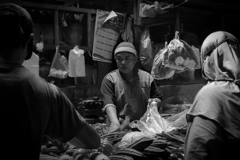 A market vendor serves customers across a produce stall with vegetables and packaged goods hanging behind him.