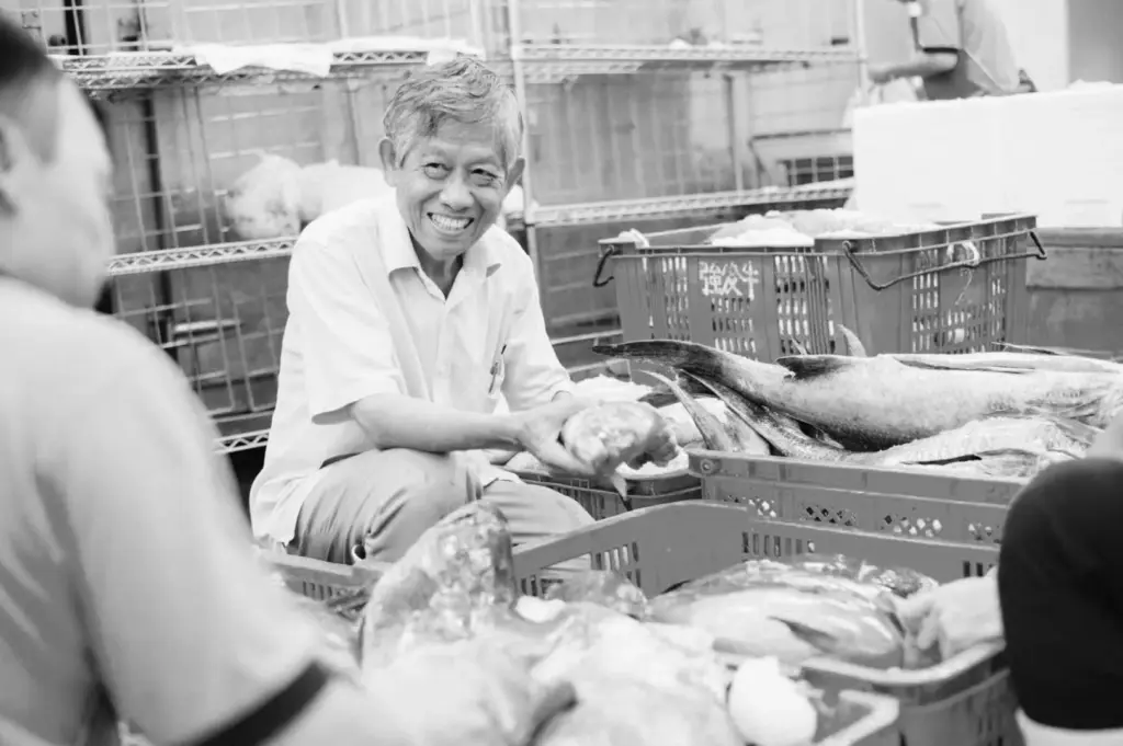 Elderly fish vendor smiling while handling fresh seafood at a fish market